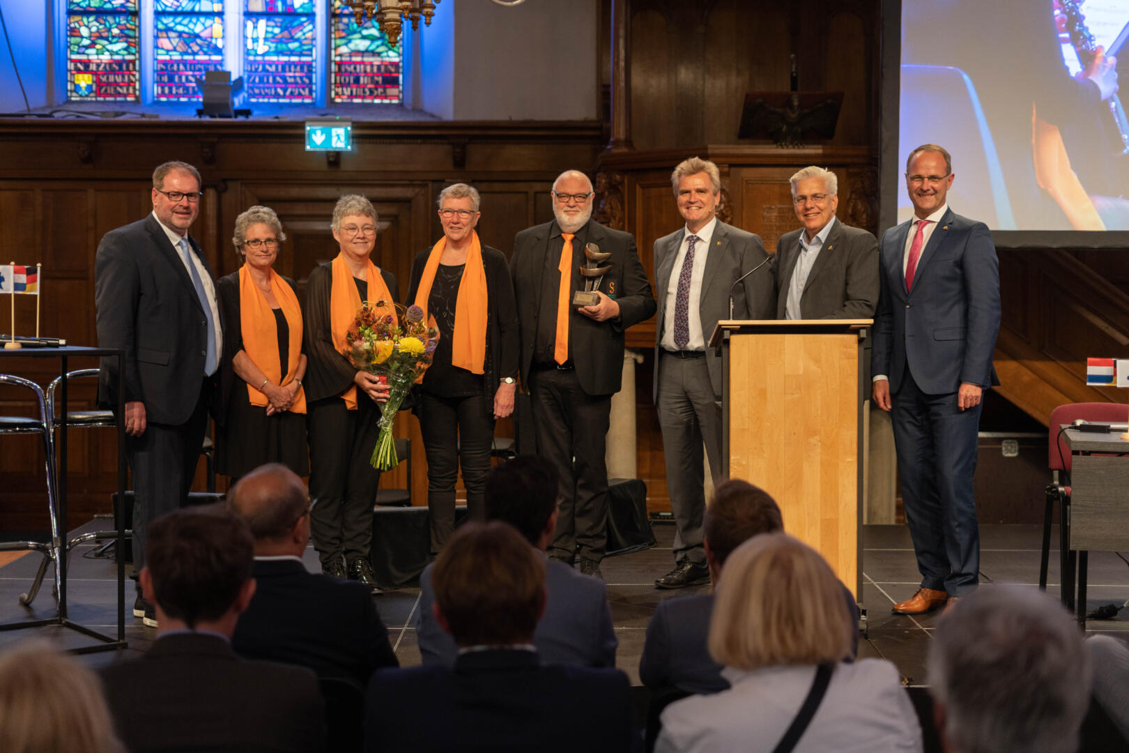 De winnaars van de People to People EUREGIO-prijs staan op het podium samen met vier bestuursleden van de EUREGIO. De winnaars houden een boeket bloemen en de prijs in hun handen.