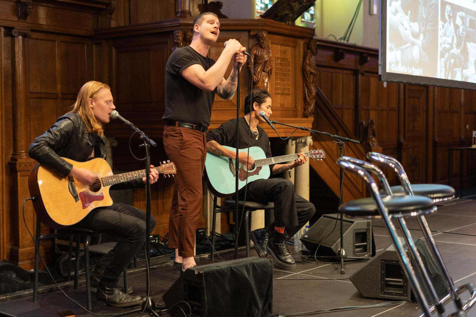 De band Ten Times A Million staat op het podium. Twee gitaristen zitten op het podium en spelen. Tussen hen staat de zanger in het microfoon te zingen.